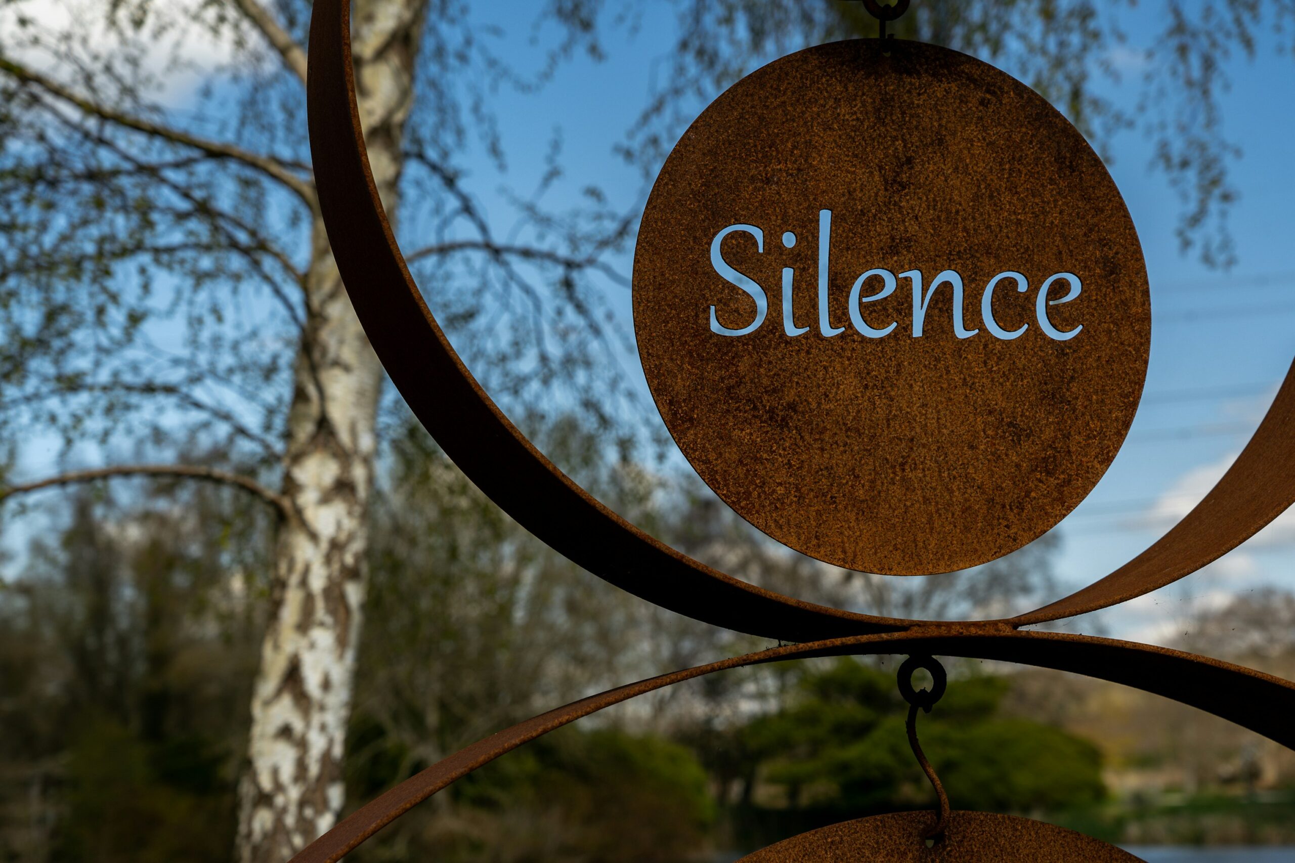 The word silence cut into an iron disk, hung as a sign from a tree with a serene blue sky in the background.